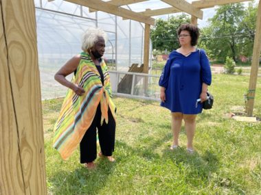 Two women stand under a pergola in a green area beside a greenhouse. The women are Vel Scott and Shani Richards.
