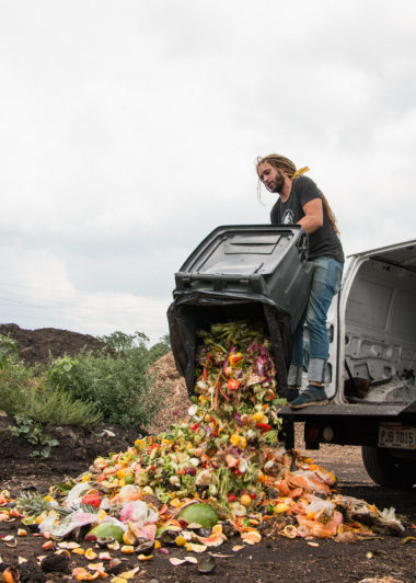 A man stands on the back of a van and unloads a dumpster full of food waste on to the ground. The good waste includes scraps like watermelon rind, tomato cores, and pineapple tops among other things.